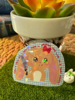 A small decorative sticker or patch featuring a cartoon rabbit character on a gingham background, placed on artificial grass next to a white planter.