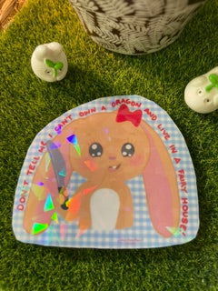A handmade paper craft plaque featuring a cartoon bunny in a fairy house, placed on artificial green grass with small decorative stones nearby.
