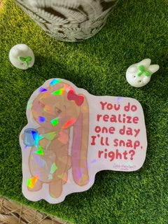 A die-cut sticker featuring a cartoon bunny with a rainbow holographic overlay and the text 'You do realize one day I'll snap right?' placed on artificial green grass next to small white decorative items.