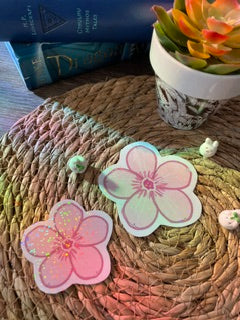 Two pink and white rainbow holographic cherry blossom sakura flower stickers or decals displayed on a woven straw mat.