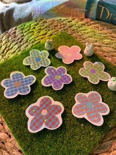 A set of flower-shaped holographic vinyl stickers with a checkered pattern in pink, purple, and blue, arranged on a green artificial grass mat.A set of six flower-shaped decorative coasters with a plaid pattern, displayed on a green mossy surface next to a woven basket.