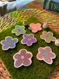 A set of flower-shaped holographic vinyl stickers with a checkered pattern in pink, purple, and blue, arranged on a green artificial grass mat.A set of six flower-shaped decorative coasters with a plaid pattern, displayed on a green mossy surface next to a woven basket.