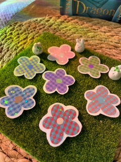 A set of flower-shaped holographic vinyl stickers with a checkered pattern in pink, purple, and blue, arranged on a green artificial grass mat.