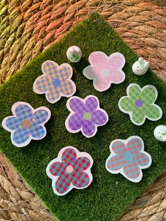 A set of flower-shaped holographic vinyl stickers with a checkered pattern in pink, purple, and blue, arranged on a green artificial grass mat.