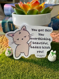 A die-cut sticker featuring a cute cartoon cat with the text 'You got this! You loverthinking, beautiful, badass bitch you!' on a white background, shown with small decorative props.
