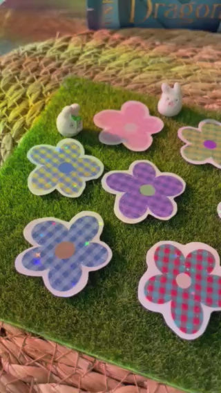 A set of flower-shaped holographic vinyl stickers with a checkered pattern in pink, purple, and blue, arranged on a green artificial grass mat.