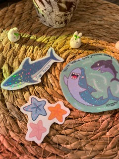 A set of cute mermaid and sea life enamel pins, including a blue shark, a purple mermaid on a green background, and three starfish in orange, blue, and pink, displayed on a woven straw mat.