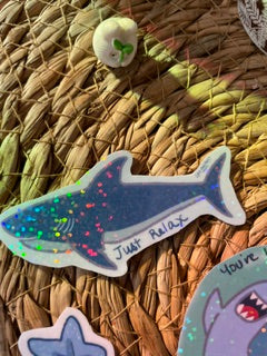 A die-cut sticker of a blue shark with colorful glittery dots and the text 'JUMP! NINJA!' on it, displayed on a woven straw hat alongside other shark-themed stickers.