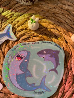 A square painted stone featuring two cartoon sharks in shades of purple and blue, displayed on a woven straw mat alongside other small decorative stones.