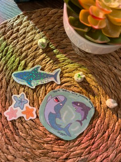 A set of three die-cut sticker charms featuring cartoon sea creatures, including a blue shark, a purple octopus, and two starfish, arranged on a woven brown mat.